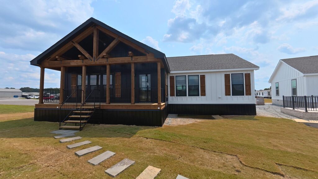 A modern, beige house with a rustic wooden porch and large windows. Stone path leads to steps; set in a grassy yard under a partly cloudy sky.