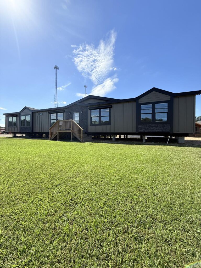 A modern, modular home with gray siding and large windows sits elevated on a sunny day. A grassy lawn stretches in front, and a blue sky with clouds is overhead.