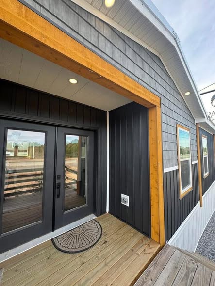 Modern tiny house exterior with dark siding, a wood-framed entrance, and glass double doors. Large windows and a welcoming mat on a wooden deck create a cozy vibe.
