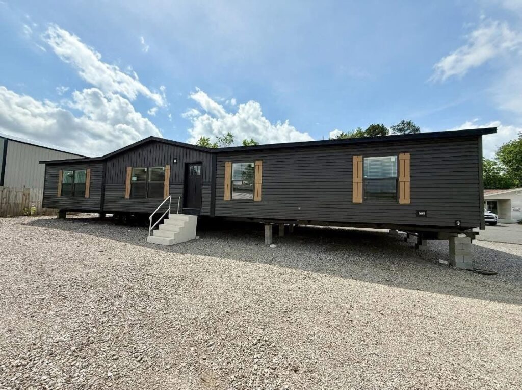 A modern, dark gray manufactured home on a gravel lot under a blue sky with clouds. The home features wooden shutters and a small staircase.