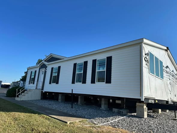 A white mobile home with black shutters is elevated on blocks, showcasing its front entrance and multiple windows. The scene is under a clear blue sky.