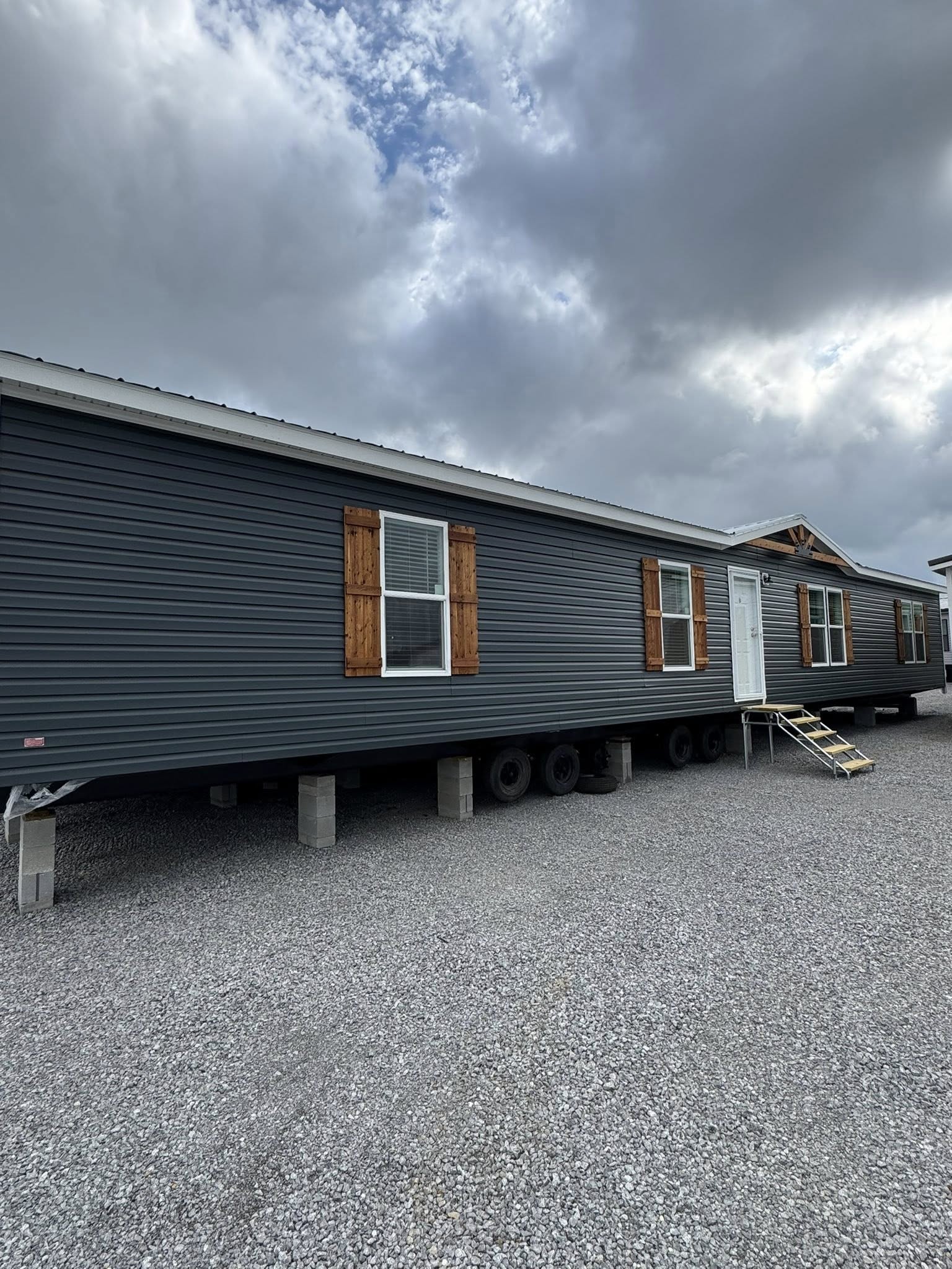 Dark blue mobile home on concrete blocks, under cloudy sky. Features white door, three windows with wooden shutters, and metal steps on gravel.
