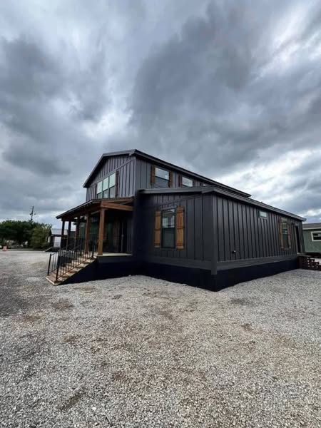 A modern, dark gray two-story house with wood accents sits on a gravel lot under an overcast sky, evoking a moody and calm atmosphere.