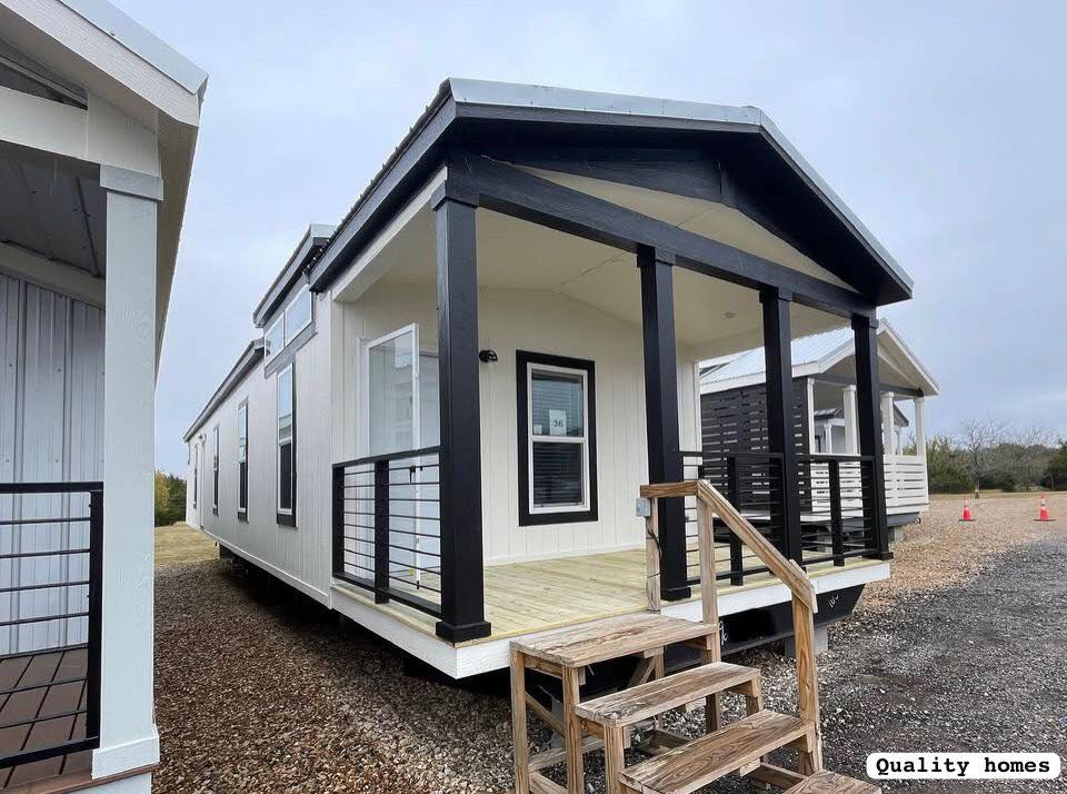 Small, modern white mobile home with black-trimmed porch and steps on gravel lot. Overcast sky, minimalistic design, welcoming atmosphere.