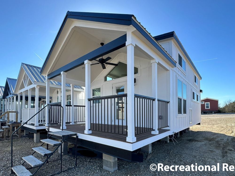 Row of modern tiny homes with blue metal roofs under a clear sky. Front house features a porch with white columns and a black railing.