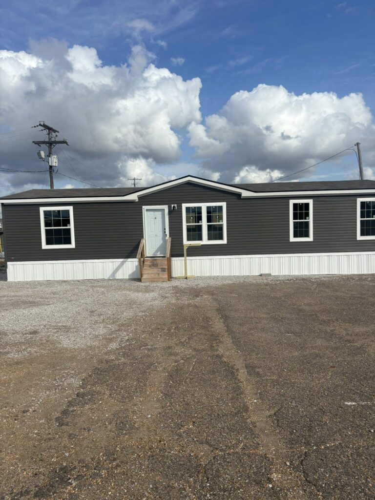 A single-story home with dark siding and a light blue door sits under a sky filled with fluffy clouds. The setting is peaceful and open.