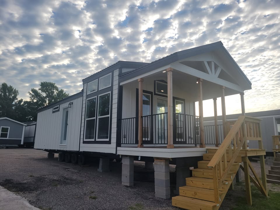 A modern tiny house with large windows and a porch sits elevated on blocks under a cloudy, bright sky. Wooden steps lead to the entrance.