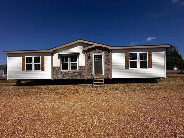 A white modular home with a gabled roof, stone accents, and brown shutters stands on a gravel lot under a clear blue sky. The front door is centered.
