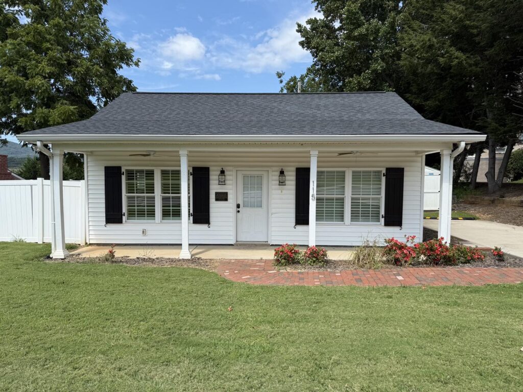 Small white house with black shutters, two columns, and a dark roof. Surrounded by green lawn and red flowers, under a bright blue sky with clouds.