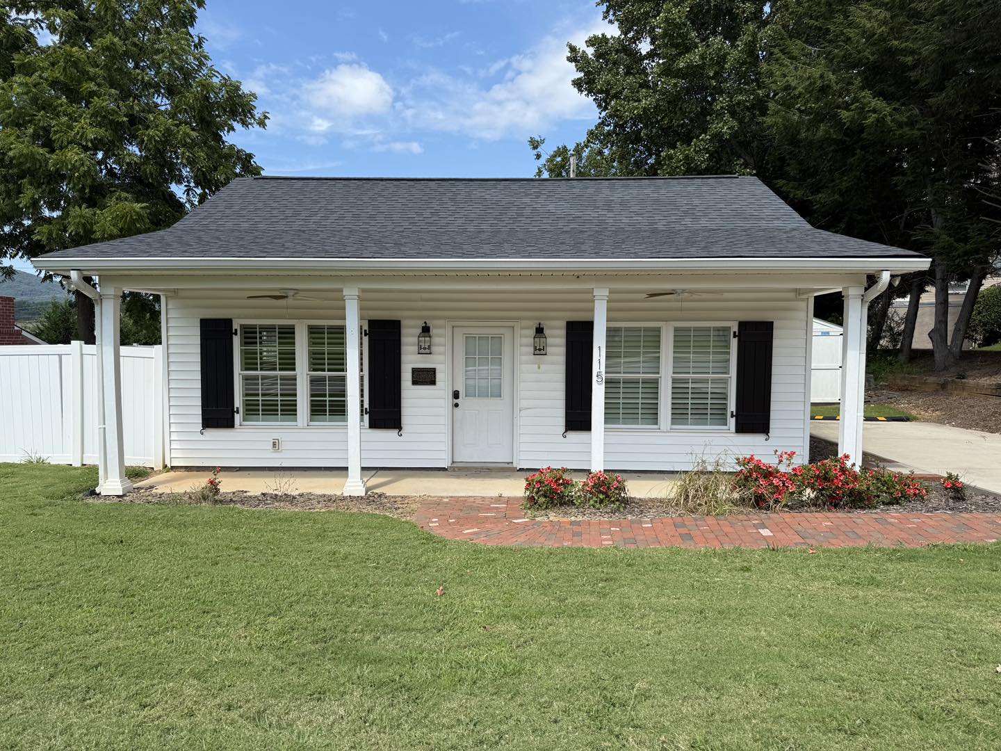 Small white house with black shutters, two columns, and a dark roof. Surrounded by green lawn and red flowers, under a bright blue sky with clouds.