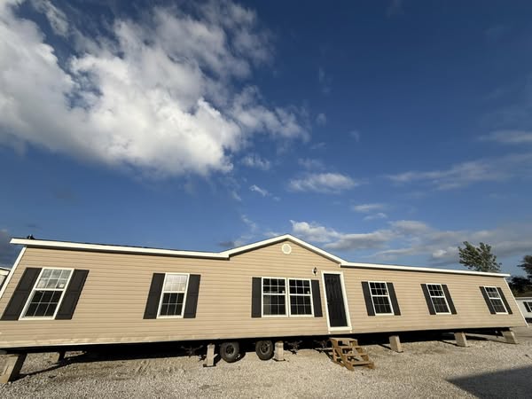 Single-story beige manufactured home on a gravel lot, with white trim and dark shutters. Set against a blue sky with scattered clouds, it conveys tranquility.