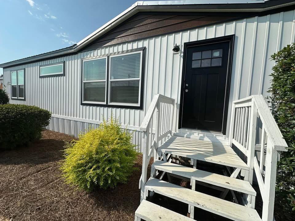 A white mobile home with a black door is framed by tidy shrubs and bushes. Wooden steps lead to the entrance, conveying a welcoming atmosphere.