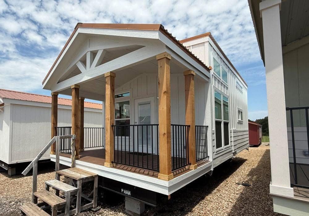 A modern tiny house with a gabled roof, wooden pillars, and a small front porch with black railings. It rests on gravel under a partly cloudy sky.
