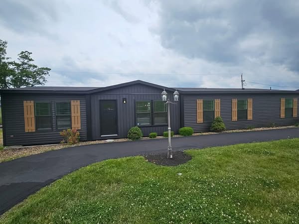 A long, dark gray modular house with tan shutters sits under a cloudy sky. A paved walkway leads to the front door, surrounded by green grass and shrubs.