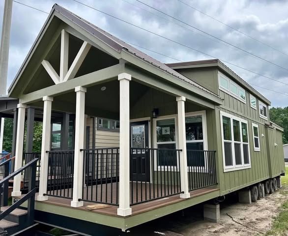 A green tiny house with white trim sits on a wheeled base, featuring a porch with railings and a gabled roof. Overcast sky adds a cozy, mobile vibe.