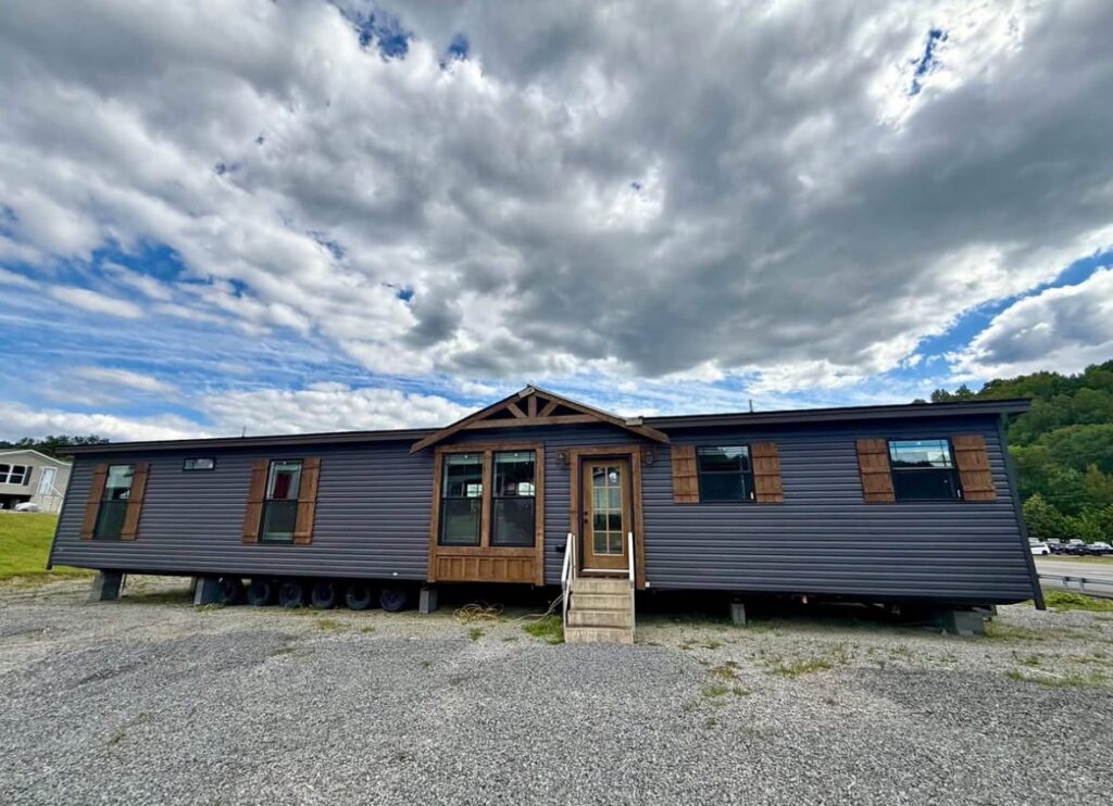 A dark gray mobile home with wooden shutters sits on a gravel lot under a partly cloudy sky. Small porch steps lead to the front door, creating a welcoming feel.