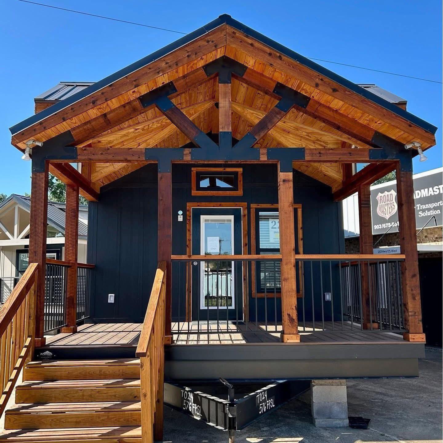 A small, modern cabin with a wooden porch and dark siding, set against a clear blue sky. The structure conveys a cozy, inviting atmosphere.