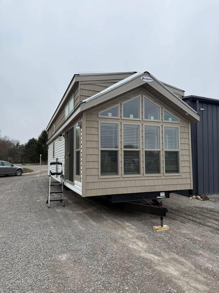A beige tiny house on wheels is parked on a gravel lot. It features large windows and a ladder leaning on one side. Overcast sky in the background.