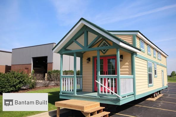A small, beige and teal tiny house with a red door sits on a paved lot. It features a wooden porch and roof accents, with a clear blue sky above.