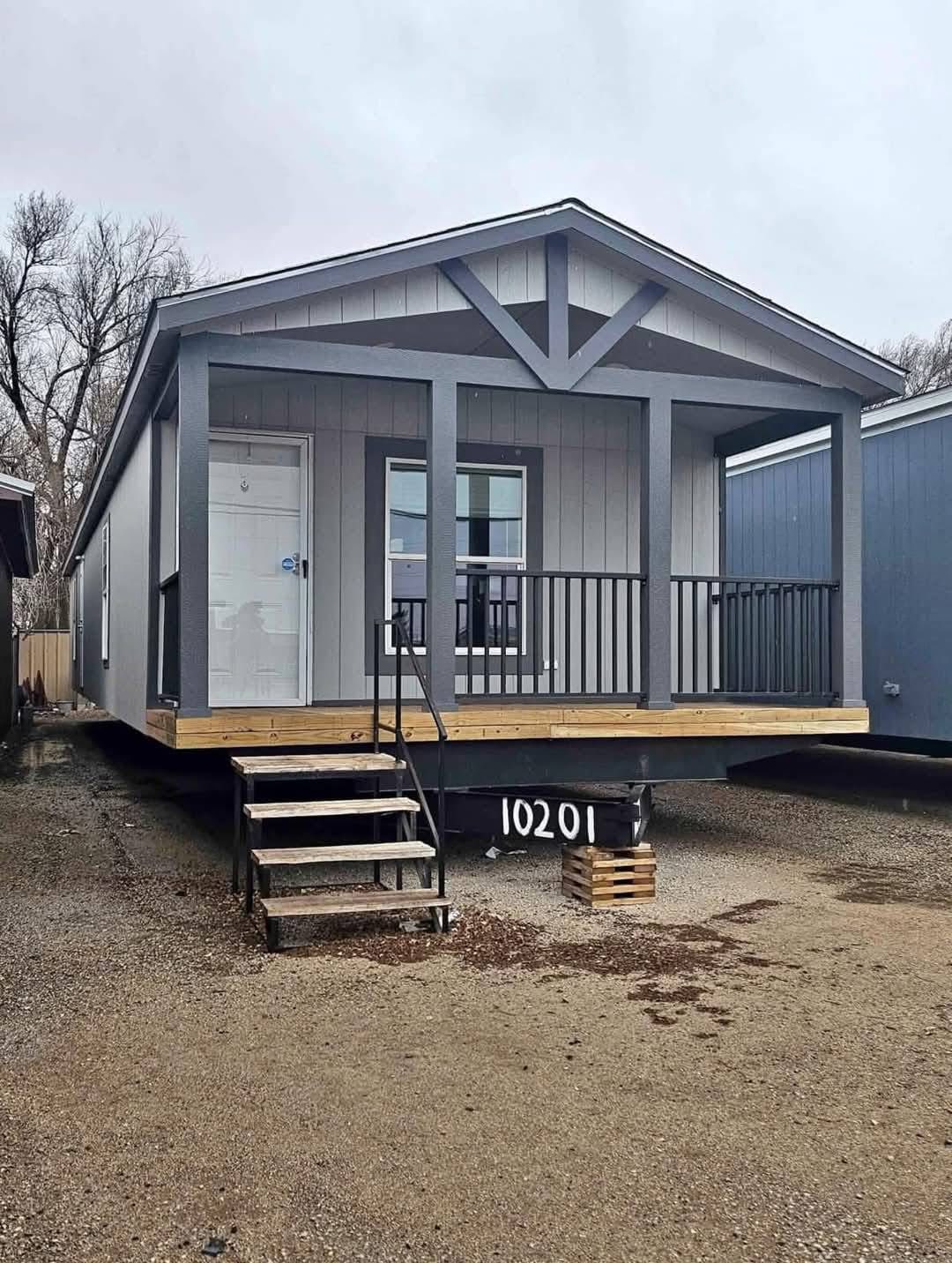 A small, gray modular home with a front porch, white door, and steps. Surrounded by gravel, the overcast sky and bare trees suggest a cold, quiet setting.