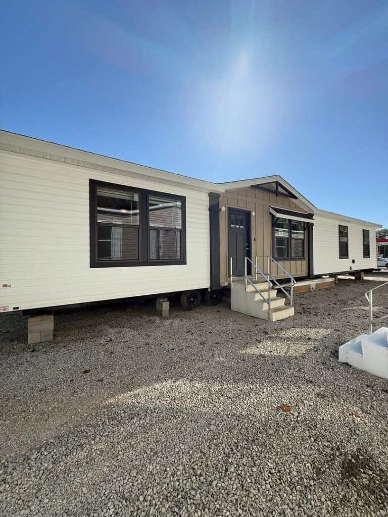 A large white mobile home with black trim sits on a gravel lot under a clear blue sky. Steps lead to the front door, conveying a sense of simplicity and brightness.