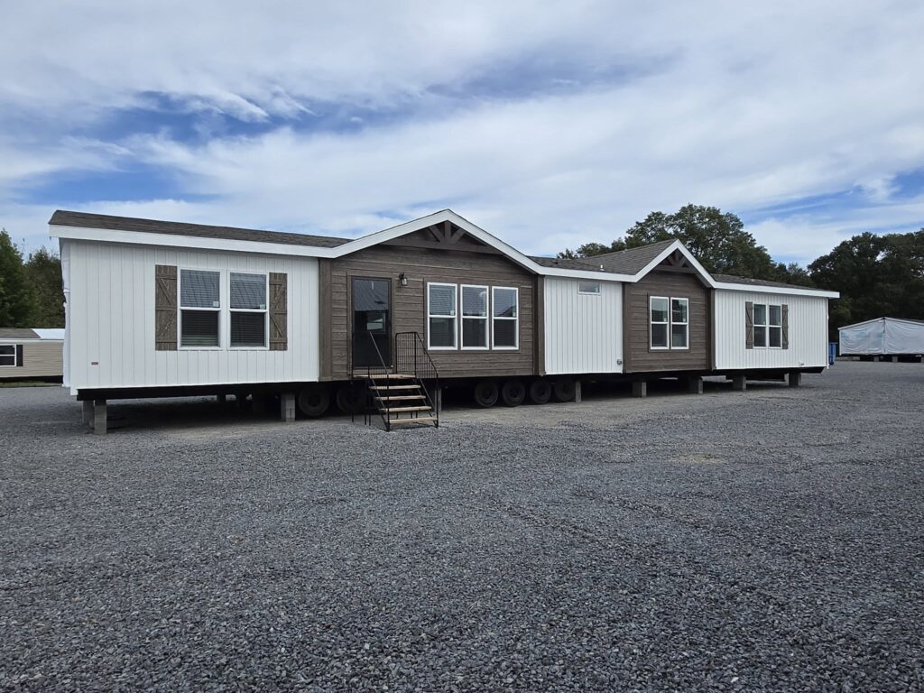 A large mobile home with brown and white siding sits on a gravel lot under a cloudy sky. There are multiple windows and a small staircase leading to the entrance.