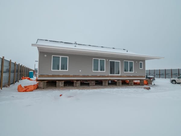 A single-story, gray modular home on stilts sits in a snowy landscape with overcast skies. Orange materials are stacked nearby, adding a touch of color.