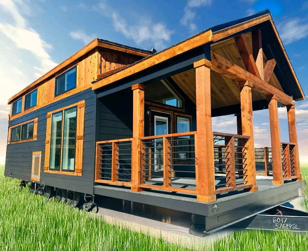 A modern tiny house with wood accents and large windows sits on a grassy field under a blue sky. The porch features a wooden railing, creating a cozy, welcoming vibe.