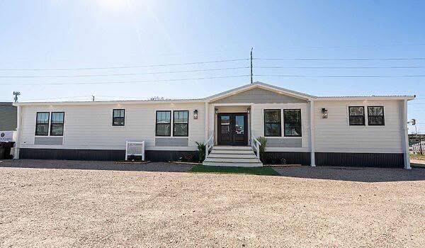 Single-story white modular home with a wide front entrance and steps. Large windows align symmetrically, and the setting is sunny with a clear blue sky.