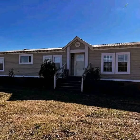 A single-story, beige manufactured home with a gable roof sits on a grassy lawn under a clear blue sky. The house has a central entrance with steps and windows on either side, flanked by shrubs.