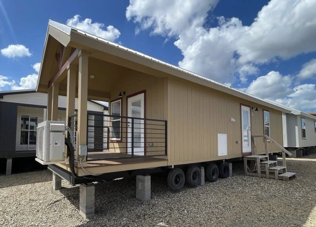 Tan mobile home on wheels with a front deck and red trim, set on gravel. The sky is blue with fluffy clouds, creating an open, welcoming feel.