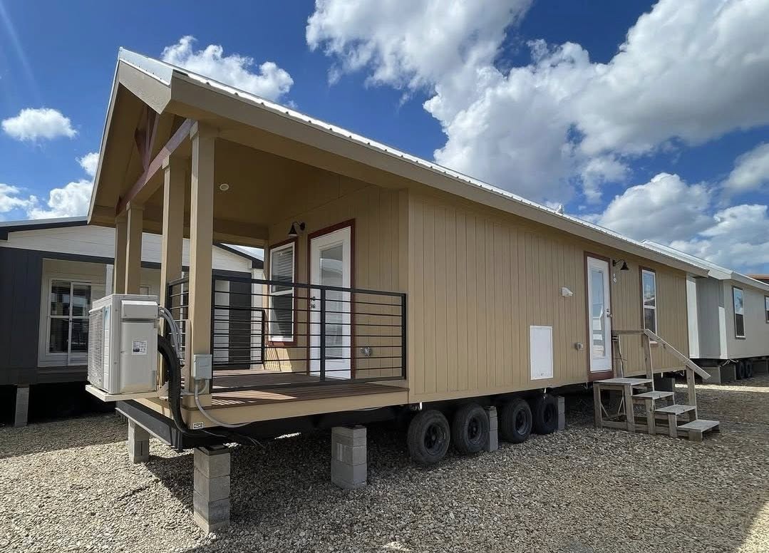Tan mobile home on wheels with a front deck and red trim, set on gravel. The sky is blue with fluffy clouds, creating an open, welcoming feel.