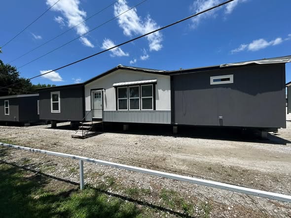 Gray and white mobile home on a gravel lot under a clear blue sky. The home has several windows and is elevated with stairs leading to the door.