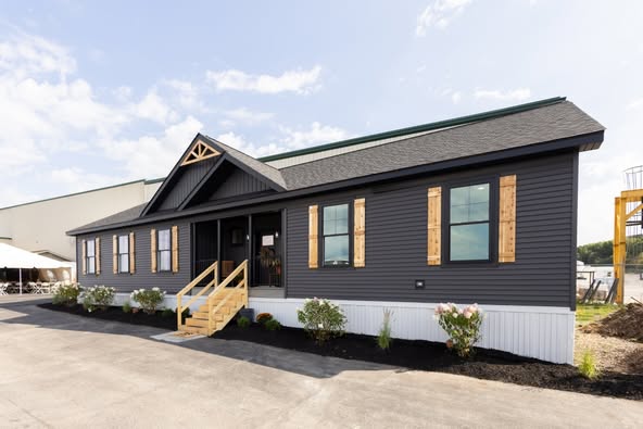 A modern, dark gray mobile home with a green roof and wooden shutters. It has a small porch with steps, surrounded by neatly planted shrubs. Clear, sunny sky.