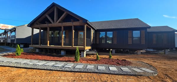 A modern, dark-toned modular home with a wooden porch and large windows. A stone path leads to the entrance, surrounded by fresh landscaping under a clear blue sky.