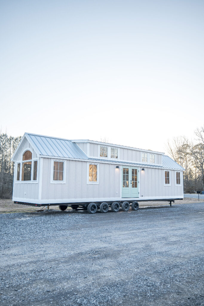 A modern white tiny house on wheels with large windows and a blue door, set against a clear sky and trees. It has a serene and minimalist vibe.