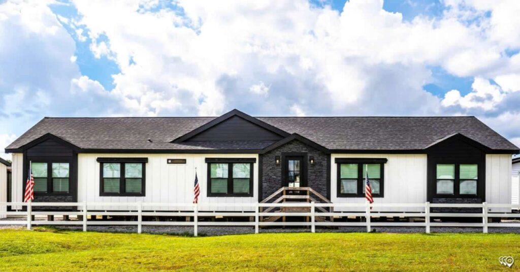 A modern, single-story house with white siding and black trim sits behind a white fence. American flags flank the entrance. The sky is partly cloudy above.