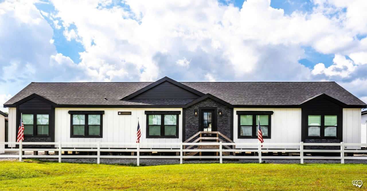 A modern, single-story house with white siding and black trim sits behind a white fence. American flags flank the entrance. The sky is partly cloudy above.