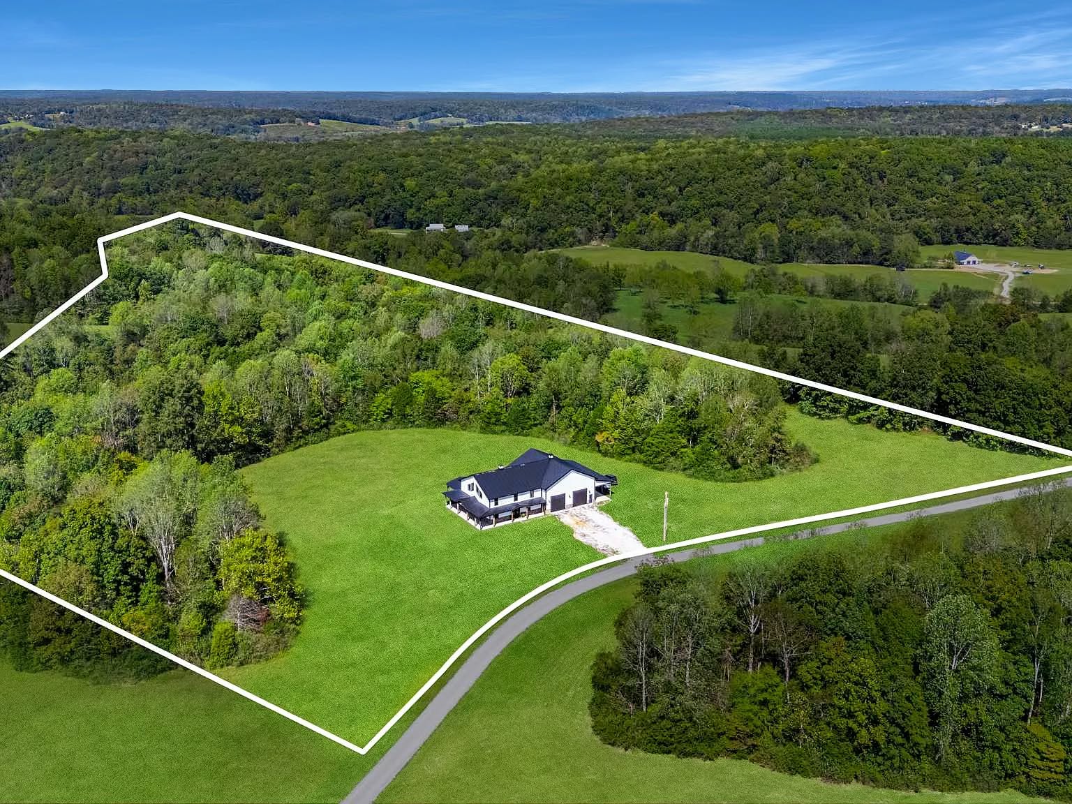 Aerial view of a large, rectangular plot of land outlined in white, featuring a modern house. The property is surrounded by dense, green trees and rolling hills under a blue sky.