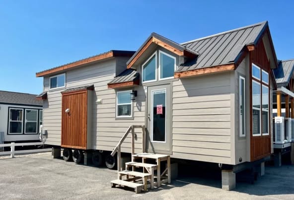 Stylish tiny house with a gabled roof, gray siding, and large windows. Wooden steps lead to the entrance. Bright, sunny day enhances the cozy feel.