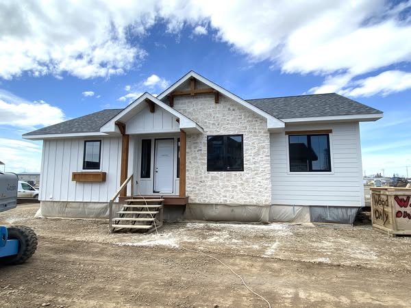 A single-story house with white siding and stone accents under a partly cloudy sky. The front features a small porch with steps and large windows.