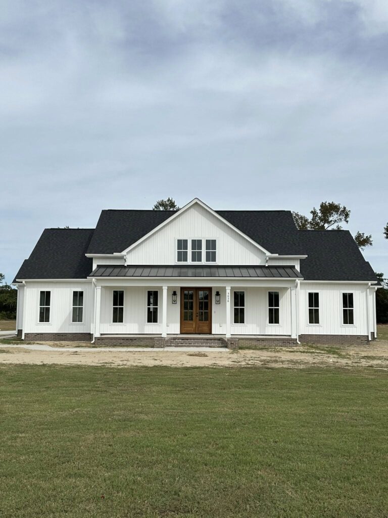 Spacious white farmhouse with a dark roof and front porch, set against a cloudy sky. Large lawn and tree-lined background create a serene setting.