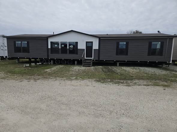 A large gray and white modular home stands on a gravel lot under a cloudy sky. It has multiple windows, a central door, and a few steps leading up.