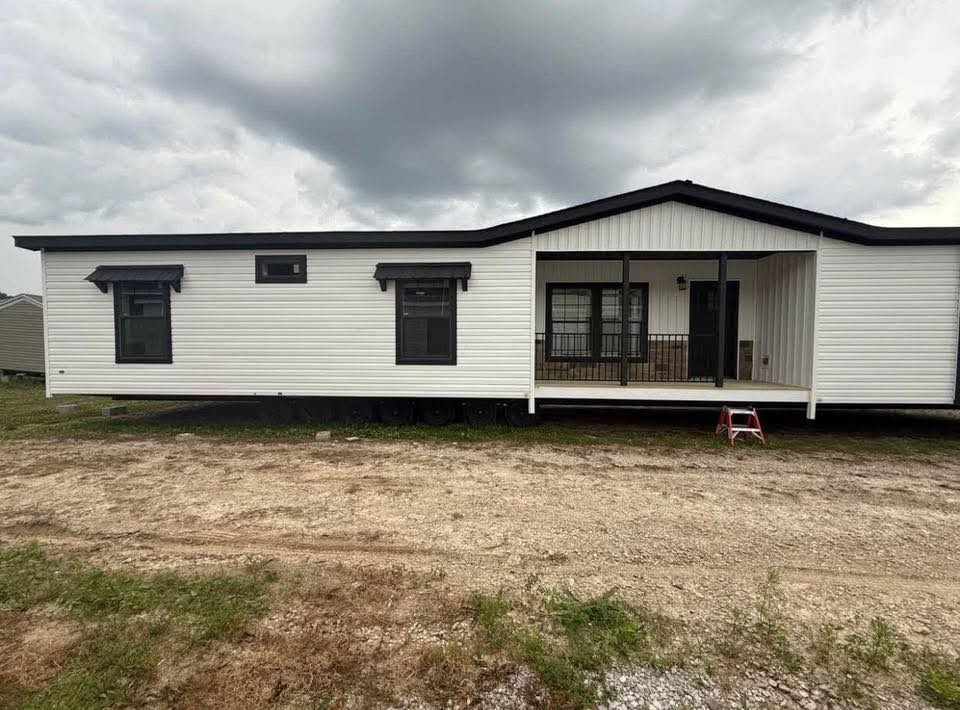 Single-story white mobile home with black trim and a front porch. Overcast sky sets a neutral tone. Positioned on a dirt and grassy lot.