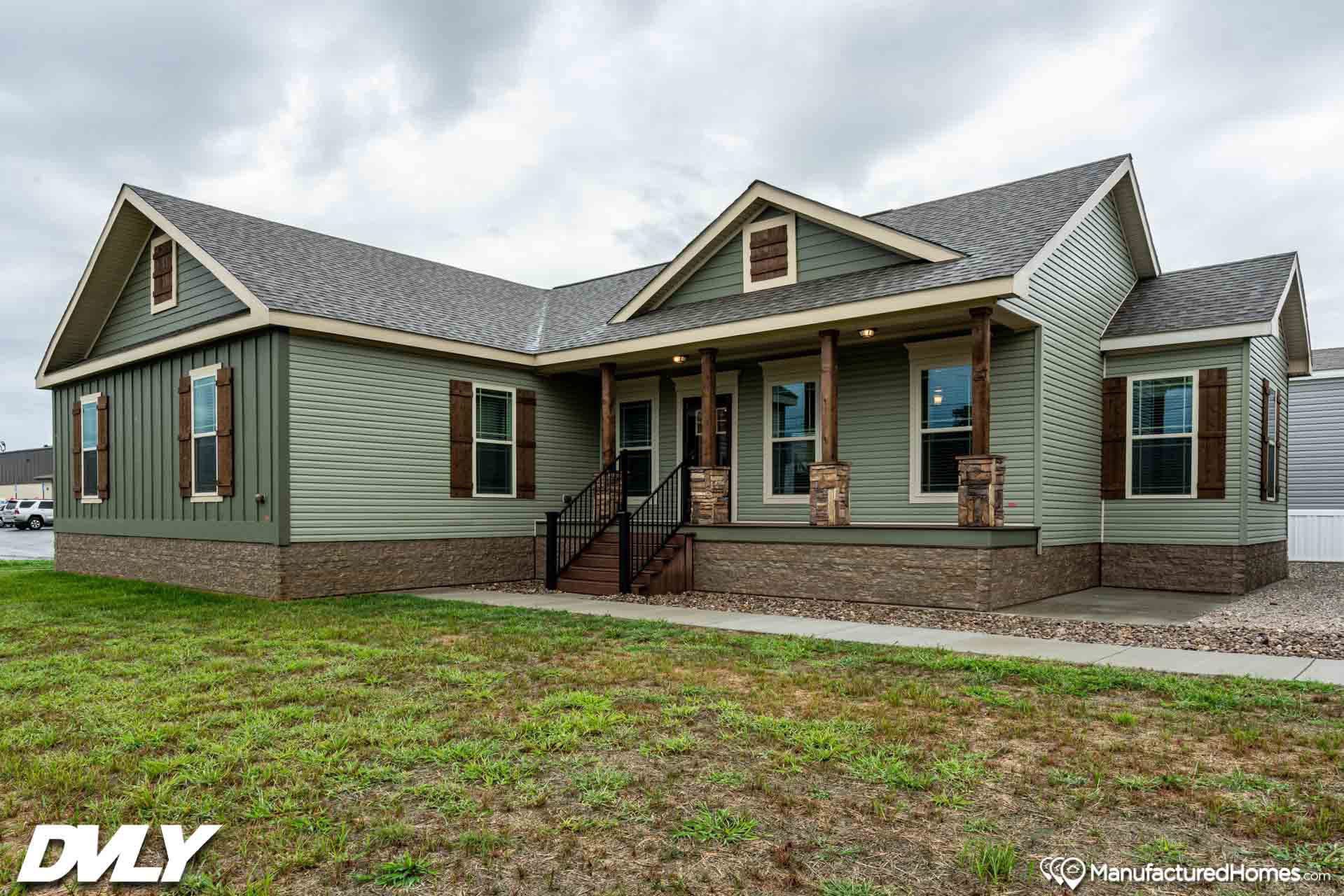 Single-story manufactured home with green siding, brown shutters, and a stone facade. It features a covered porch with wooden columns on a cloudy day.