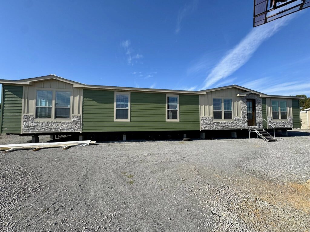 A long, green mobile home with stone accents sits on a gravel lot under a clear blue sky. Three steps lead to a door, conveying a sense of simplicity.