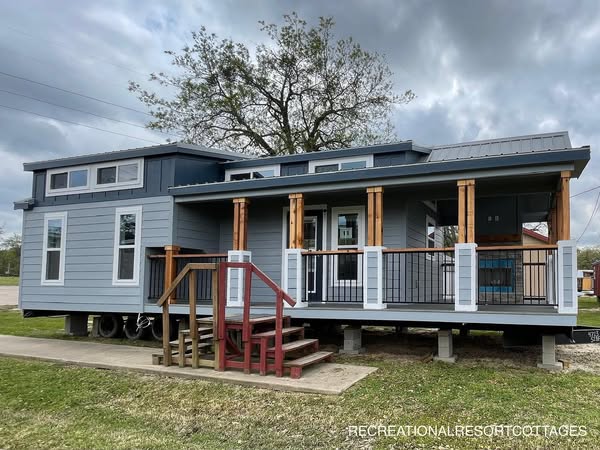 Stylish tiny house with blue siding and large windows, set against a cloudy sky. Features a wooden porch with red steps, evoking a cozy, modern feel.