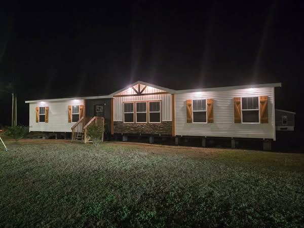 A well-lit single-story modular home with white siding and wooden accents is seen at night. Warm exterior lights create a cozy and welcoming ambiance.