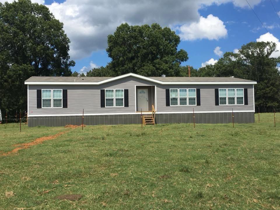 A single-story gray mobile home with white trim and black shutters sits in a grassy field. Large trees and a partly cloudy sky provide a serene backdrop.