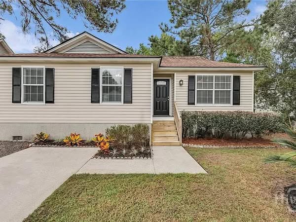 Single-story beige house with black shutters, large windows, and a black front door. Neatly maintained lawn, colorful flower beds, and shrubs.